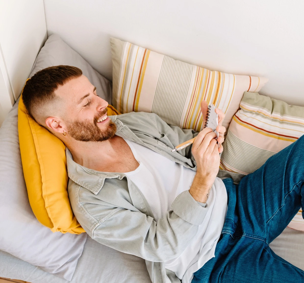 A young man lies on a gray sofa, resting on a yellow pillow, writing in a notebook, smiling