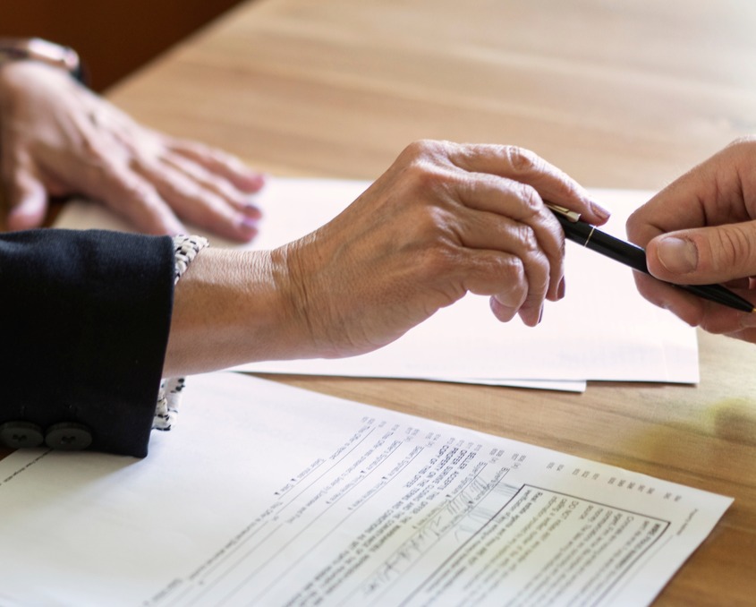 Documents are placed on the wooden table, a pen is passed from one hand to another