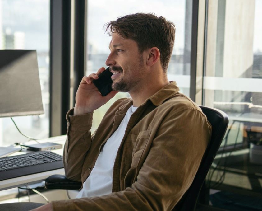 A man sits at a desk in an office, talking on a mobile phone. A computer monitor and a laptop are on the desk