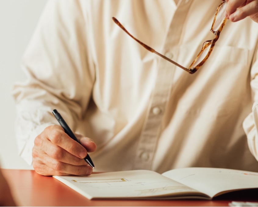 A person in a white shirt sitting at a table, holding a pen in one hand and glasses in the other, preparing to sign a document.