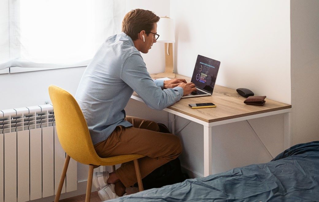 A young man sits on a yellow chair at a table with a laptop, wearing blue shirt and brown pants
