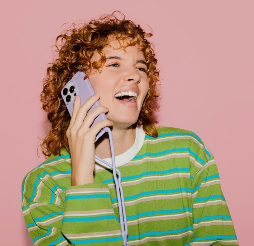 A girl with ginger hair and green striped blouse in the pink background is talking on the phone and laughing