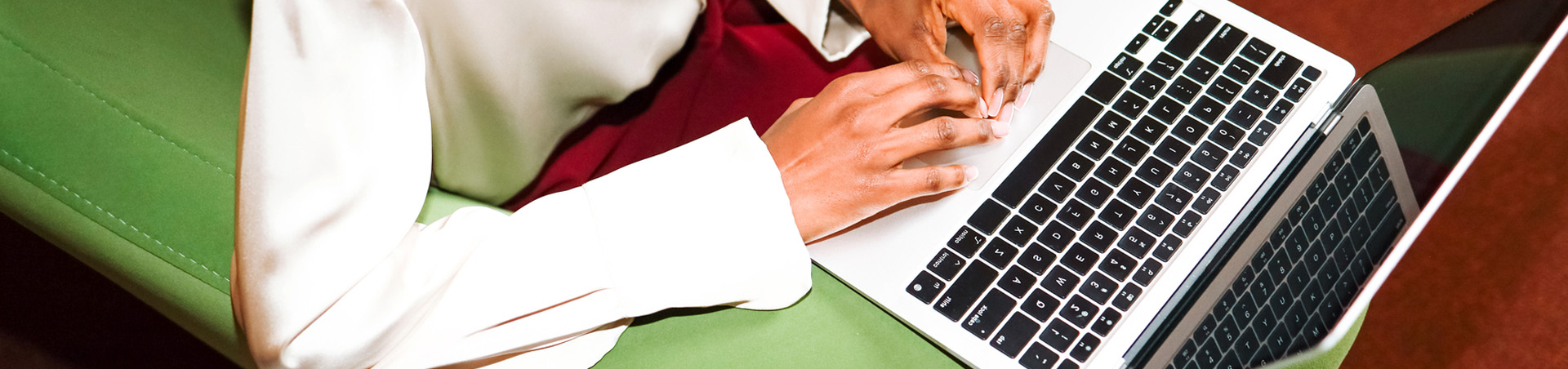 A woman in red pants and white shirt sits on a green beanbag, working on a laptop